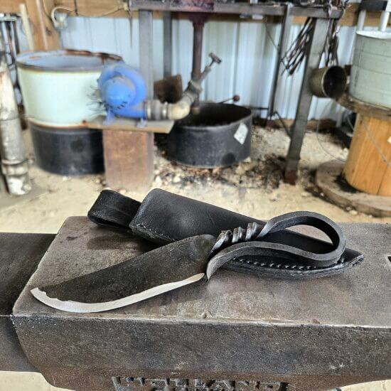 Hand-forged one-piece Iron Age knife with a twisted steel handle and loop, resting on a Holland anvil next to a black leather sheath, with a glowing forge in the background.