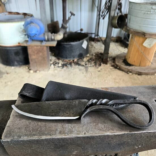 Hand-forged one-piece Iron Age knife with a clean finish, twisted steel handle and loop, resting on a Holland anvil next to a black leather sheath, with a glowing forge in the background.
