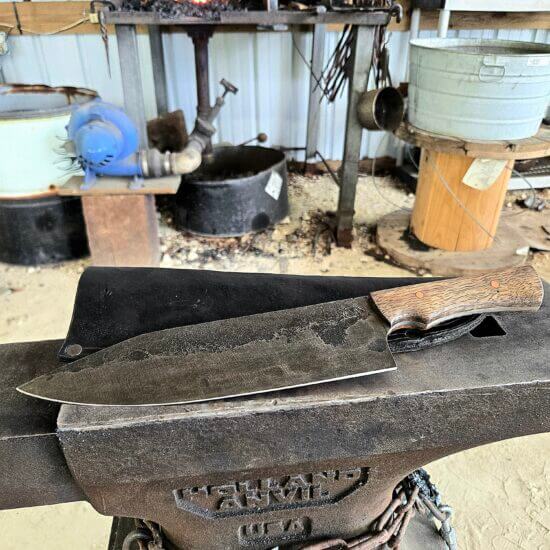 Large hand-forged 13 1/5" Gyuto kitchen knife with a dark stonewashed blade and a fiery-grained Mango wood handle, resting on a Holland anvil with a glowing forge in the background.