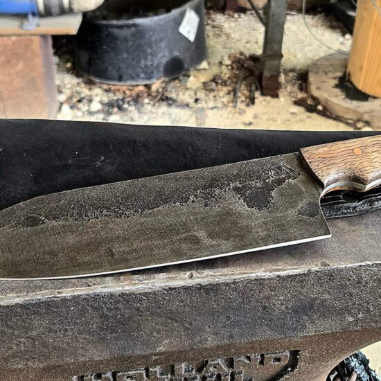 Large hand-forged 13 1/5" Gyuto kitchen knife with a dark stonewashed blade and a fiery-grained Mango wood handle, resting on a Holland anvil with a glowing forge in the background.