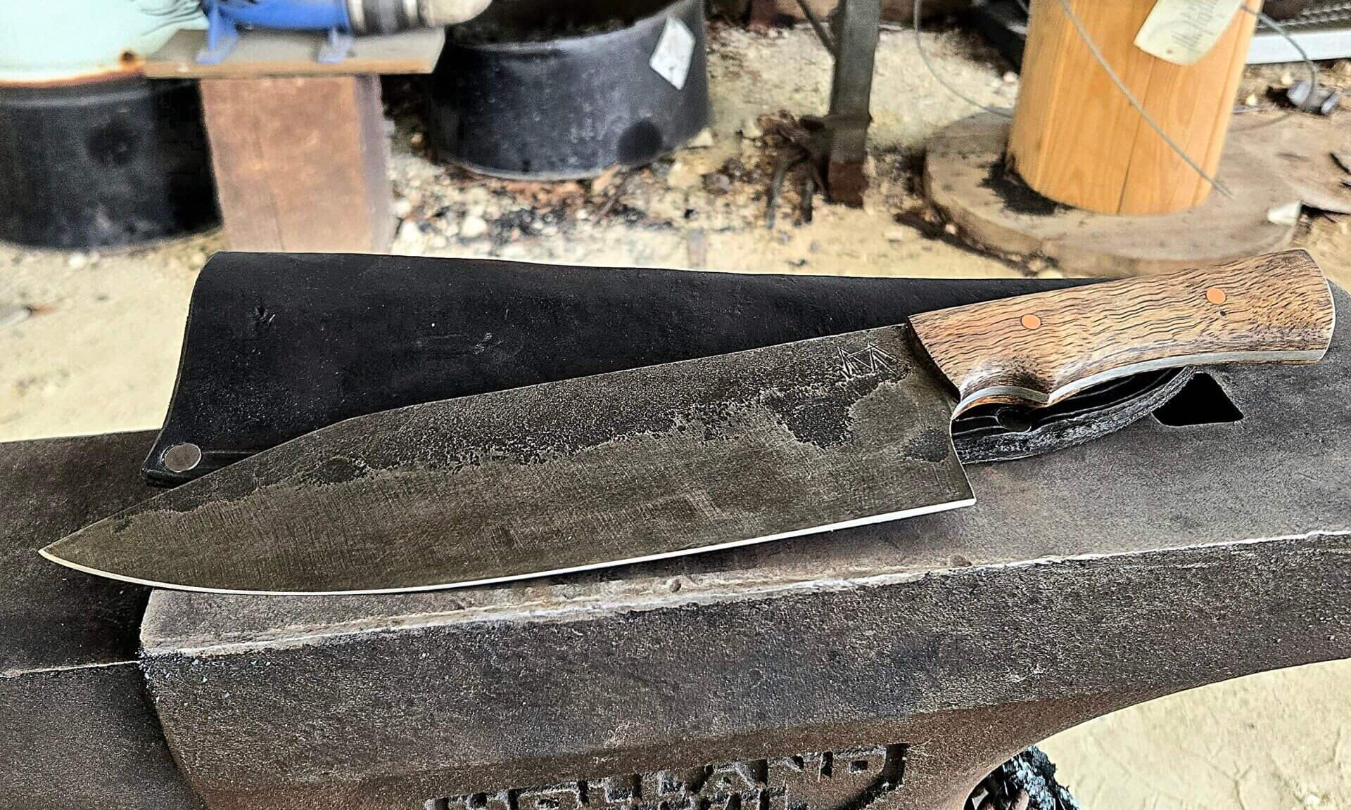 Large hand-forged 13 1/5" Gyuto kitchen knife with a dark stonewashed blade and a fiery-grained Mango wood handle, resting on a Holland anvil with a glowing forge in the background.